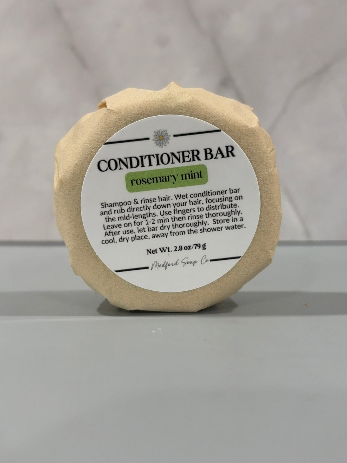 A single round conditioner bar wrapped in beige paper packaging with a white label reading 'CONDITIONER BAR' and 'rosemary mint,' photographed on a light gray marble-like surface.