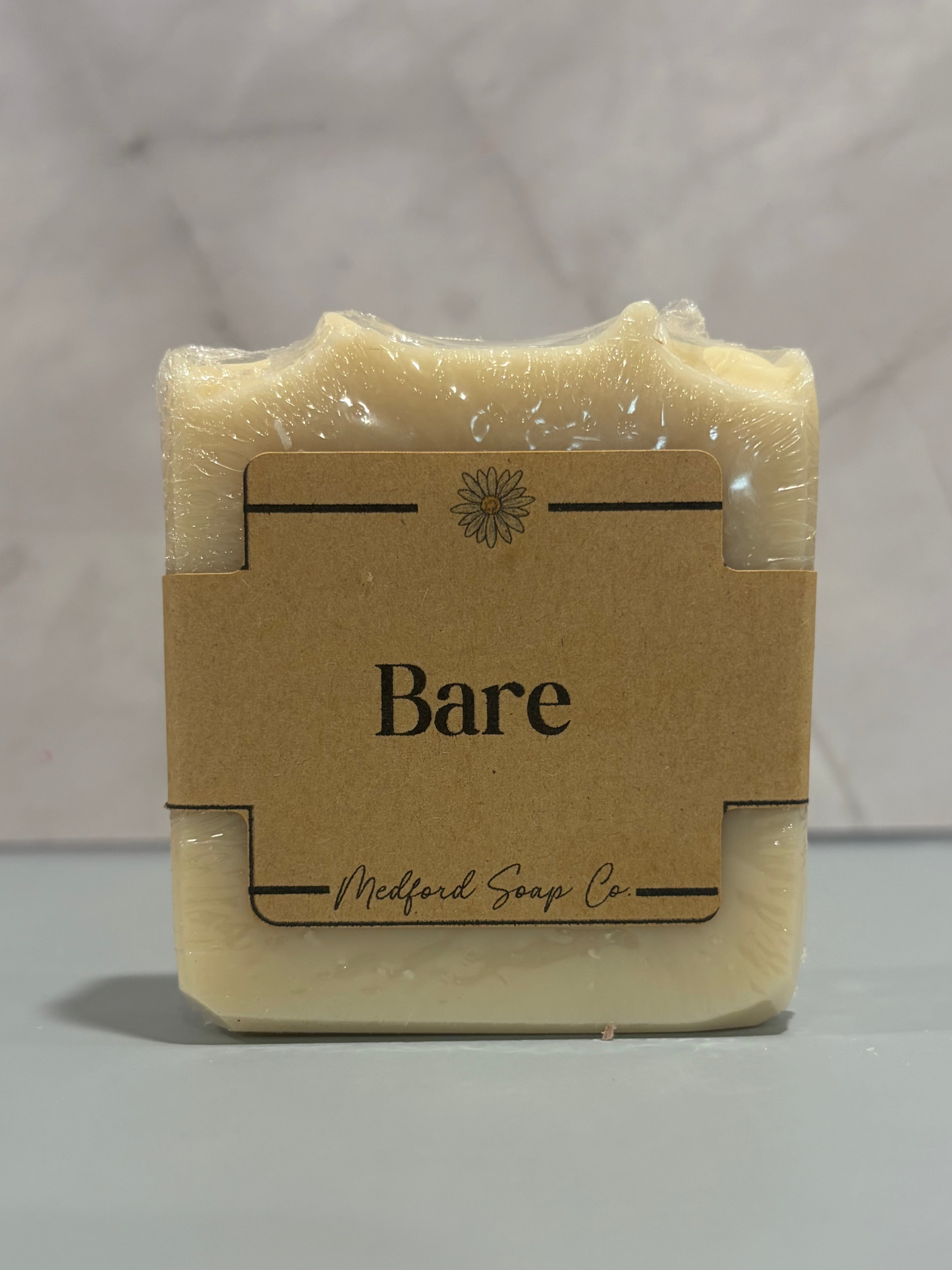 A rectangular bar of pale cream-colored soap wrapped in clear plastic and secured with a brown kraft paper label that reads 'Bare' and 'Midford Soap Co.' on a light gray marble-patterned background.