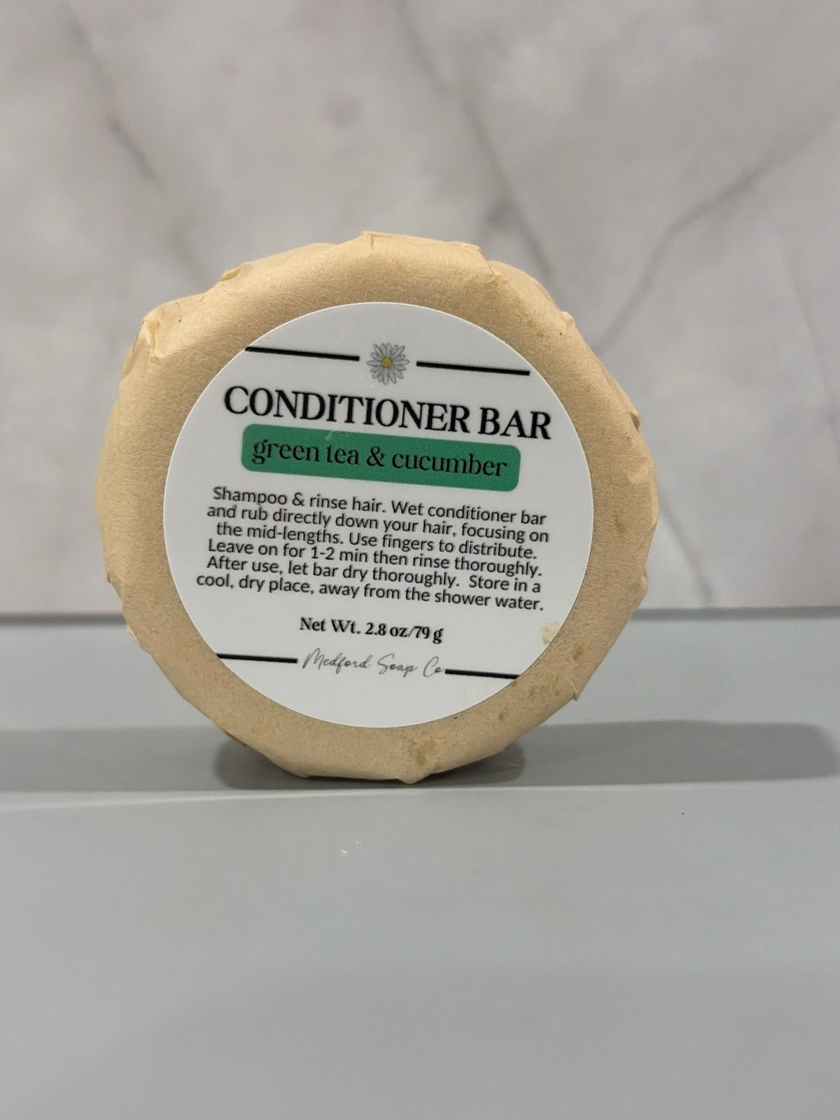 A round natural conditioner bar with a white paper label reading 'CONDITIONER BAR green tea & cucumber', set against a light gray marble background.