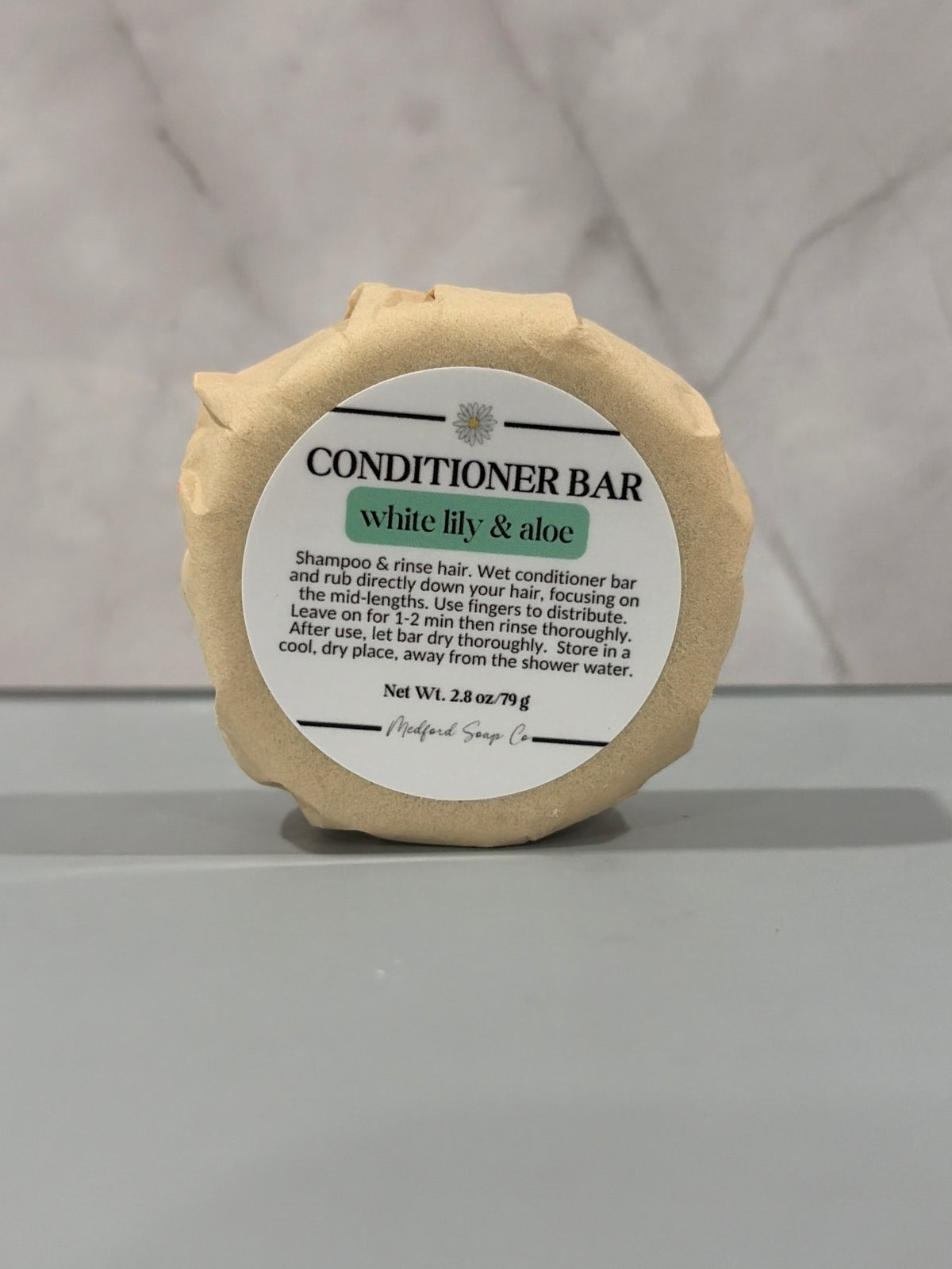 A single round conditioner bar wrapped in kraft paper with a white paper label reading 'CONDITIONER BAR white lily & aloe' and 'Medford Soap Co.' on a gray surface against a light marble-patterned background.