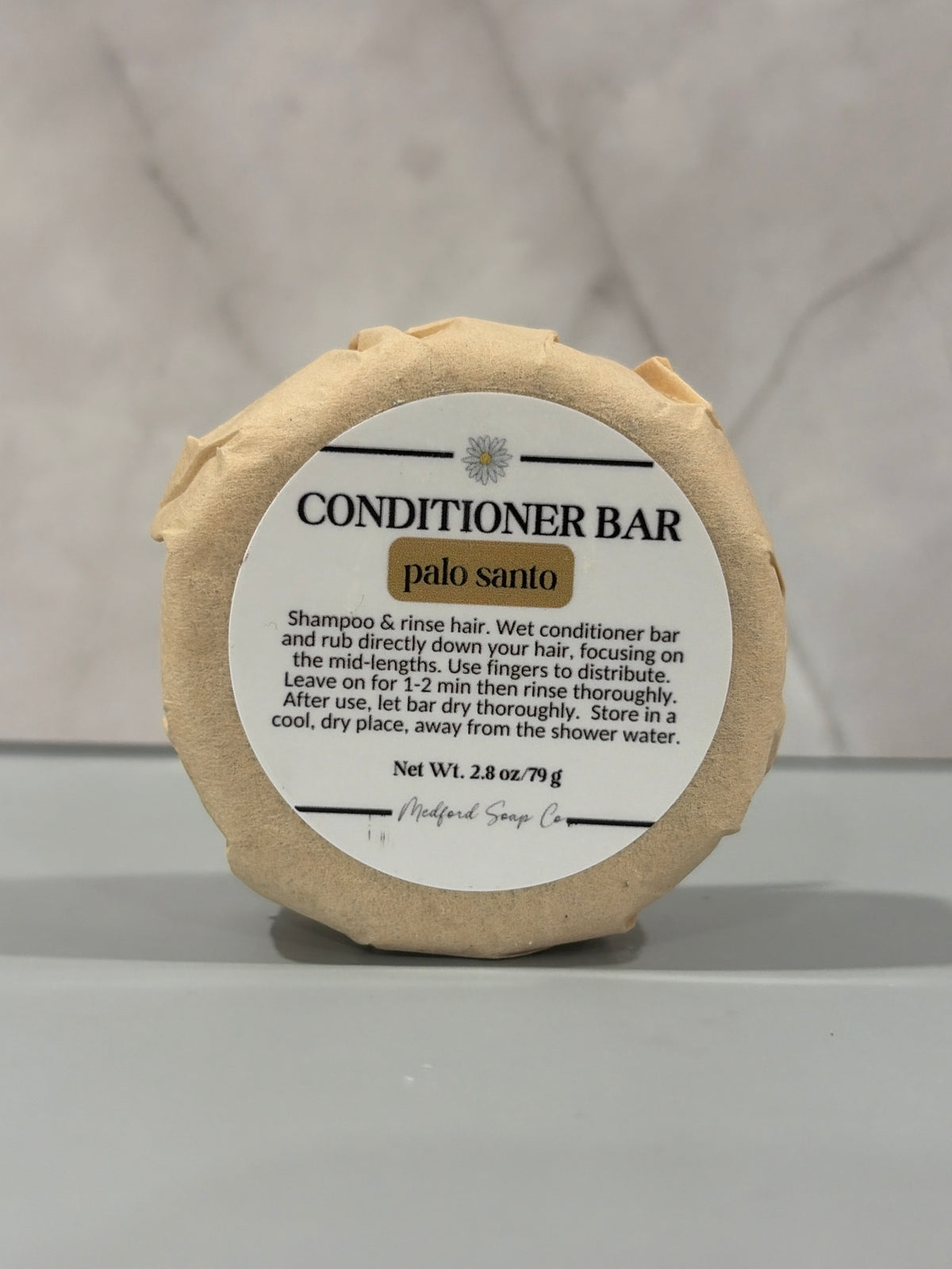 A round conditioner bar wrapped in beige paper with a white paper label that reads 'CONDITIONER BAR palo santo' on a light gray marble-like background.