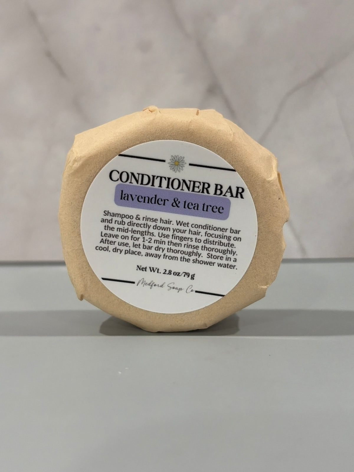 A round, conditioner bar wrapped in kraft paper with a white paper label that reads 'CONDITIONER BAR lavender & tea tree', set against a light gray marble background.